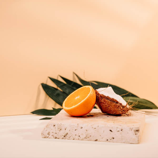 Aufgeschnittene Orangenhälfte und halbe Kokosnuss liegen auf einer beigen Fliese, Palmwedel im Hintergrund