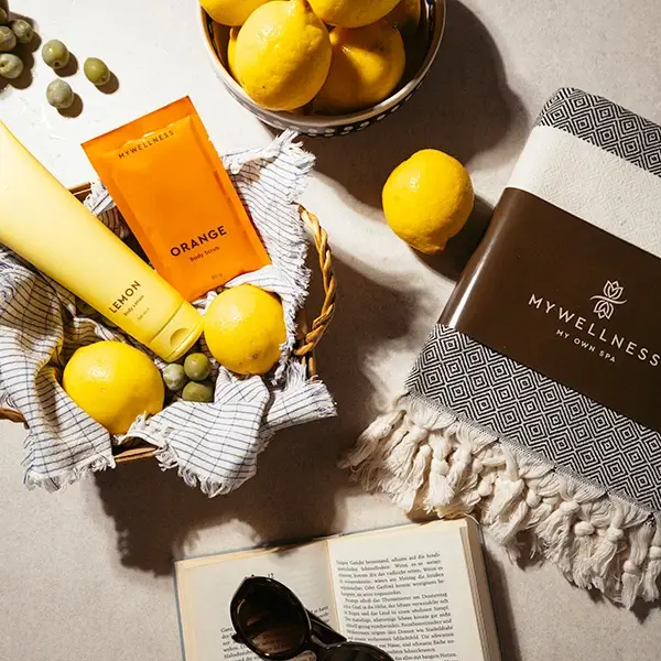 MyWellness Lemon Body Lotion und Orange Body Scrub in einem Weidenkorb mit Zitronen, MyWellness Hamam-Tuch, aufgeschlagenem Buch und Sonnenbrille