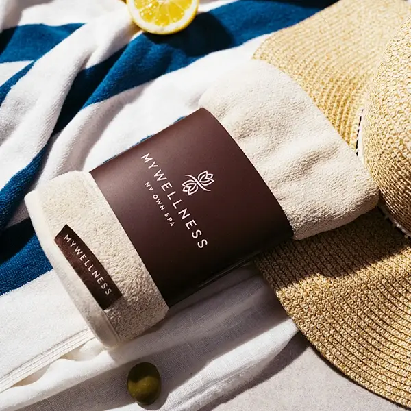 MyWellness Handtuch in cremeweiß mit Banderole auf blau-weißem Strandtuch neben Strohhut und Zitrone