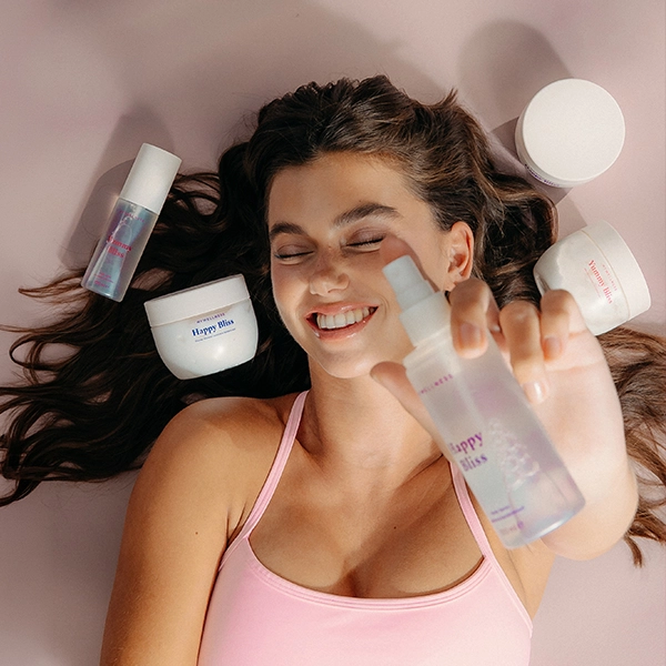 Junges Model liegt auf rosafarbenen Boden und streckt das Yummy Bliss Bodyspray in Richtung Kamera. Um sie herum liegen verschiedene Happy Bliss und Yummy Bliss Produkte von MyWellness
