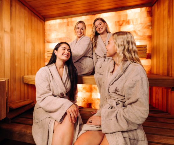 Vier Frauen in MyWellness-Bademänteln unterhalten sich lachend in der Sauna mit Salzsteinwand