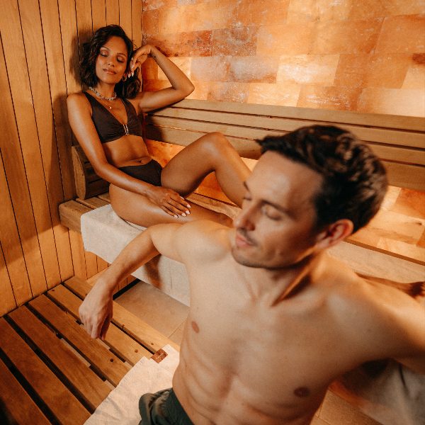 Ein Mann und eine Frau entspannen sich gemeinsam in einer holzvertäfelten Sauna mit warmem Salzsteinambiente. Beide haben die Augen geschlossen und wirken ruhig und erholt.